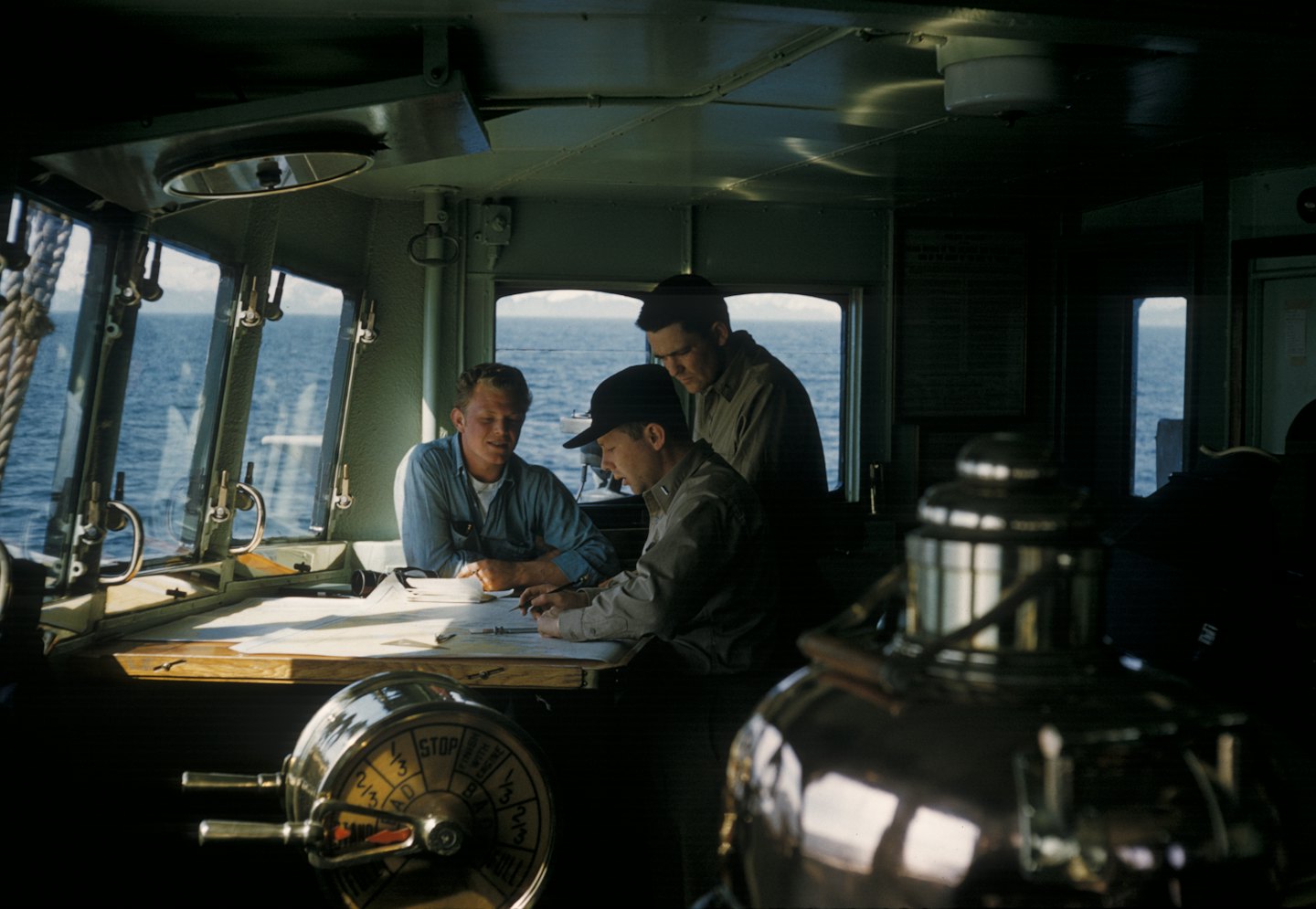 Tre uomini esaminano carte nautiche sul ponte di una nave con l'oceano all'esterno.