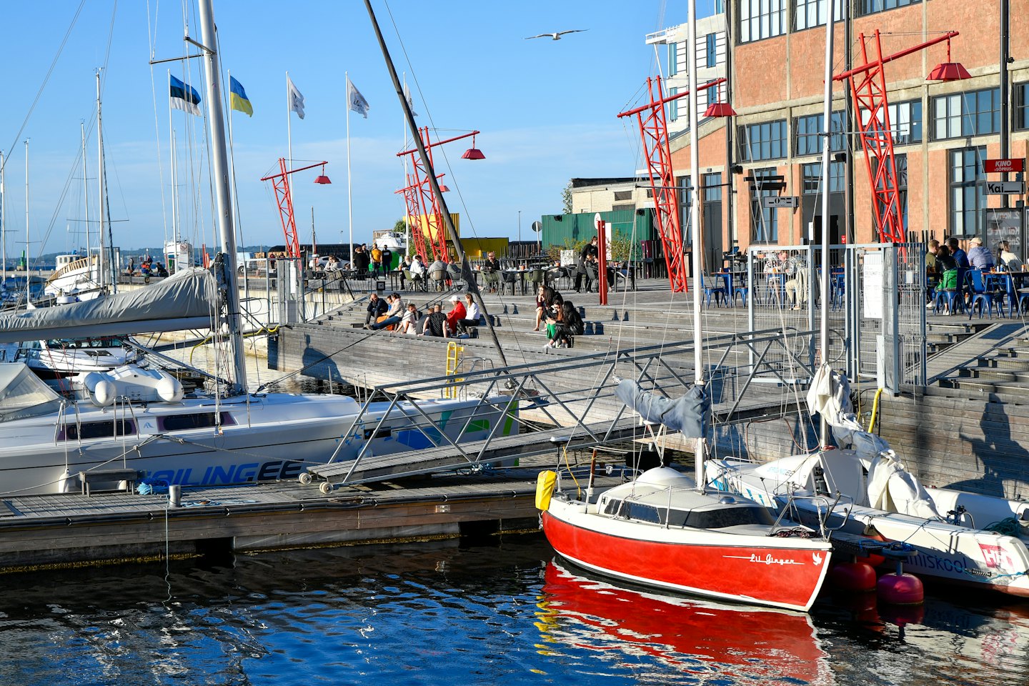 Barche a vela ormeggiate in un porto turistico con persone che cenano sul molo.